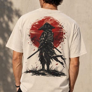 May include: An off-white short-sleeve t-shirt featuring a large graphic on the back. The design depicts a black silhouette of a samurai wearing a conical hat and holding two swords, standing before a large, textured red sun with ink-like splatters.