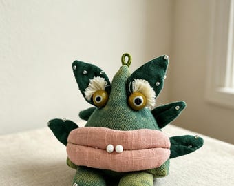 May include: A plush creature made of dark green textured fabric with a large, horizontal, light pink mouth featuring two small white bead teeth. It has two round yellow eyes with white fringe accents, small pointed limbs, and silver bead embellishments.