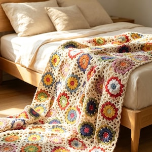 Könnte beinhalten: Eine Häkeldecke mit einem sich wiederholenden Muster aus kreisförmigen Blumenmotiven in verschiedenen Farben wie Rot, Gelb, Blau, Rosa und Orange, die durch ein cremefarbenes Gittermuster verbunden sind. Die Decke liegt auf einem Bett mit heller Bettwäsche.