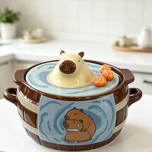May include: A ceramic pot with a lid featuring a sculpted capybara head and three small orange citrus fruits. The pot has a brown barrel-like design with a light blue panel displaying a graphic of two capybaras, one holding a small white knife.