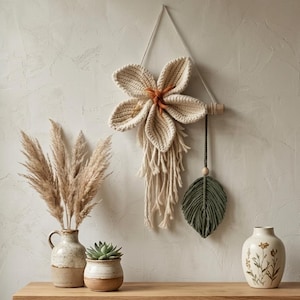 Op de afbeelding: Een wandhanger met een grote, crèmekleurige gebreide bloem met oranje vezels in het midden, opgehangen aan een houten stok. Onder de bloem hangen crèmekleurige franjes naast een donkergroen macramé blad aan een apart koord.
