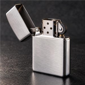 Può includere: Un accendino in metallo argento spazzolato con corpo rettangolare e coperchio incernierato aperto. Il meccanismo interno presenta una rotella circolare per la pietra focaia, una base color ottone e un camino con fori di ventilazione.