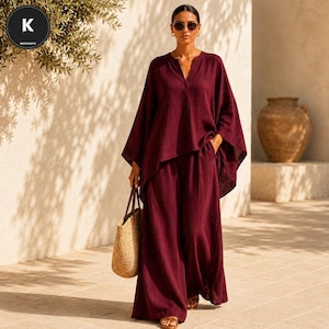 May include: A deep burgundy two-piece set consisting of a loose-fitting, long-sleeved tunic top with a V-neckline and matching wide-leg trousers. The fabric has a textured, crinkled appearance. A woven straw tote bag is carried at the side.