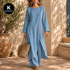 May include: A light blue long-sleeved tunic dress featuring a side-slit design and a vertical row of brown buttons extending from the neckline down the front. The garment is paired with matching wide-leg trousers and a woven straw tote bag.