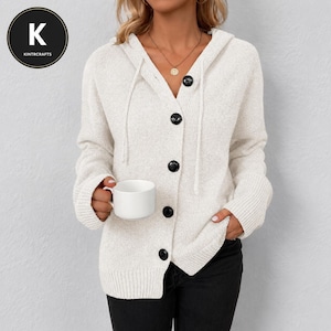 May include: A cream-colored knit hooded cardigan featuring a V-neckline, long sleeves with ribbed cuffs, and a front button closure with five large black buttons. The garment includes a matching drawstring hood and is worn over black pants.
