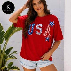 Puede incluir: Una camiseta roja de manga corta con las letras USA en el pecho. La U es azul con estrellas blancas, la S tiene un estampado floral y la A presenta rayas rojas y blancas con estrellas azules en los puntos superior e inferior de la letra.