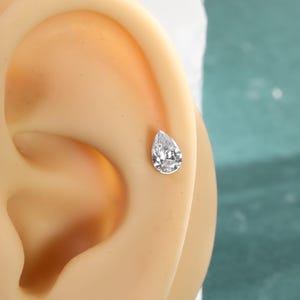 May include: A single teardrop-shaped stud earring featuring a clear, faceted gemstone set in a metallic frame. The earring is positioned on the upper cartilage area of a beige-colored ear model, showcasing the jewelry's reflective surface.