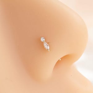May include: A gold nose stud featuring a small round clear gemstone positioned above a marquise-cut clear gemstone. The jewelry is set in a thin metal post and is displayed on a neutral-toned anatomical model of a human nose.