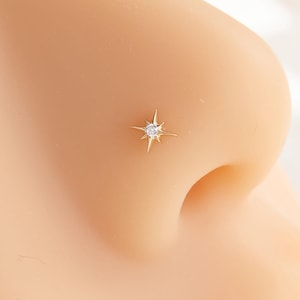 May include: A gold-toned nose stud featuring a small, central clear stone set within a four-pointed starburst design. The jewellery is positioned on the side of a beige-coloured anatomical model of a human nose, showcasing the star-shaped metal detail.