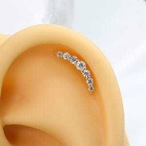 Puede incluir: Un pendiente de cartílago curvo en tono dorado con una hilera de seis piedras transparentes de corte redondo en tamaños graduados. La joya está colocada en el borde superior de un modelo de oreja beige, formando un arco de piedras brillantes.