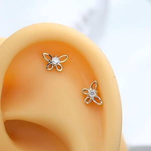 May include: Two small gold-toned butterfly-shaped earrings featuring open-work wings and a single clear round gemstone at the center of each. These stud earrings are positioned on the upper cartilage area of a beige ear model.