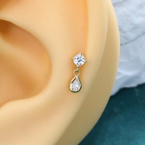 May include: A gold-toned stud earring featuring a round, clear, faceted stone at the top and a dangling, pear-shaped, clear, faceted stone drop. The earring is secured in the cartilage area of a beige-colored, synthetic ear model.
