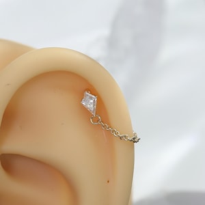 Peut inclure: Une boucle d'oreille de cartilage dorée composée d'un petit bijou transparent en forme de cerf-volant, relié par une fine chaîne métallique à un second clou, conçue pour être portée le long du bord supérieur du cartilage de l'oreille.