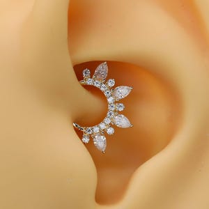 Puede incluir: Una pieza de joyería para piercing de oreja en forma de media luna dorada, con una serie de pequeñas piedras transparentes de corte pera y redondo dispuestas en un patrón de rayos de sol a lo largo del borde exterior de la banda metálica.