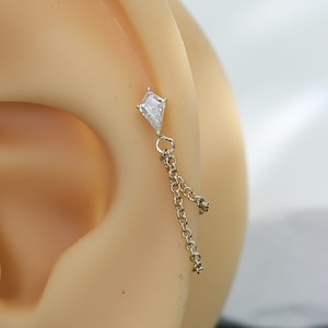 May include: A silver-toned ear piercing jewelry piece featuring a kite-shaped clear gemstone stud connected to two delicate dangling chains. The jewelry is inserted into the upper cartilage area of a neutral-toned synthetic ear model.