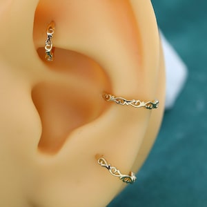 May include: Three gold hoop earrings with an intricate, repeating vine-like pattern along the outer surface. The hoops are displayed on a beige ear model, with one earring in the upper cartilage, one in the conch, and one in the lower lobe position.