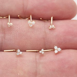 May include: Five gold-toned nose studs, each featuring a small cluster of three clear, round-cut stones arranged in a triangular pattern. The studs have a curved L-shaped post design, with the decorative stone cluster positioned at the end of the post.