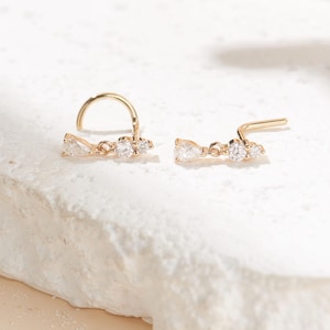 May include: Two gold nose studs featuring a delicate horizontal bar design. Each stud is accented with three small, clear, faceted stones, including a pear-shaped stone at one end and two round stones, set in a polished gold metal frame.