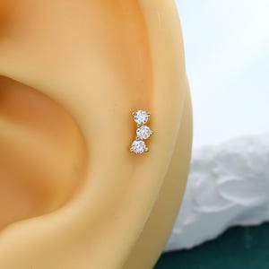 May include: A gold-toned ear stud featuring three small, clear, round-cut stones arranged in a vertical line. The jewelry is positioned on the cartilage area of a neutral-toned ear model, showcasing a delicate and simple design with a polished metal finish.