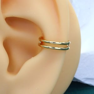 May include: A gold-toned ear cuff featuring a double-band design. The jewellery piece consists of two parallel, smooth, thin circular wires that wrap around the cartilage of the ear, creating a layered appearance with a polished, reflective finish.