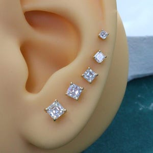 May include: A set of five gold-tone stud earrings featuring square-cut clear stones in a graduated size arrangement, displayed along the outer edge of an ear model, with each stone secured by a four-prong setting in a classic minimalist design.