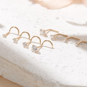May include: A collection of six gold nose studs, each featuring a small, clear, round-cut gemstone set in a prong setting. The studs have a curved, L-shaped post design, arranged in two rows on a textured, off-white, irregular stone surface.