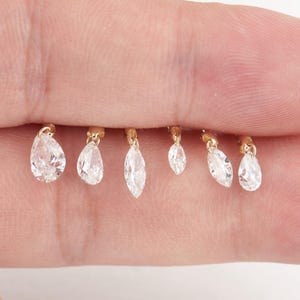 May include: A collection of six small gold-toned charms, each featuring a single clear, faceted gemstone. The charms vary in shape, including pear and marquise cuts, and are suspended from small gold loops, creating a delicate jewelry component.