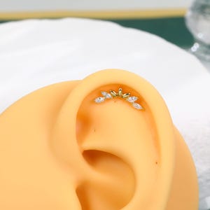 May include: A gold-toned ear cartilage stud featuring a curved, fan-like arrangement of five marquise-cut clear stones and two small gold metal accents. The jewelry is positioned along the upper curve of a synthetic ear model.