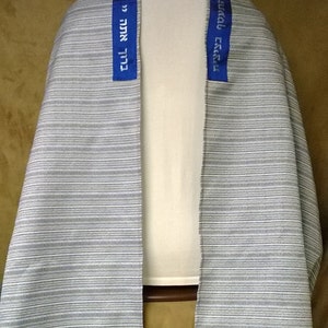 May include: A striped, gray and white tallit, a Jewish prayer shawl, draped over a form. Blue bands with Hebrew text are sewn onto the shawl. Blue squares are at the bottom.