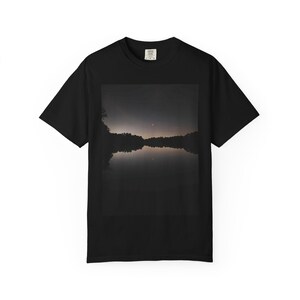 以下が含まれることがあります： 胸元に長方形のグラフィックプリントが施された黒の半袖Tシャツ。プリントには、薄暗い地平線の下、静かな水面に木々のシルエットが映り込む、夕暮れ時の暗く穏やかな風景が描かれています。