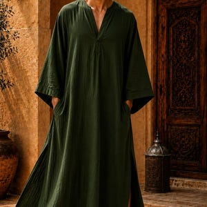 May include: A dark olive green floor-length kaftan featuring a V-neckline, wide three-quarter length sleeves, and side slits. The garment is constructed with a textured fabric and includes two side pockets, paired with dark brown leather sandals.