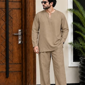 Può includere: Una camicia a maniche lunghe color beige con scollo a V e pantaloni abbinati dal taglio ampio. L'outfit è completato da sandali in pelle marrone e occhiali da sole scuri. Il completo presenta una tinta neutra e un design semplice e pulito.