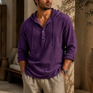 May include: A long-sleeved purple hooded shirt featuring a textured fabric, a partial button-down placket with two visible buttons, and matching drawstring cords at the neckline. The shirt is paired with light beige trousers with a straight-leg cut.