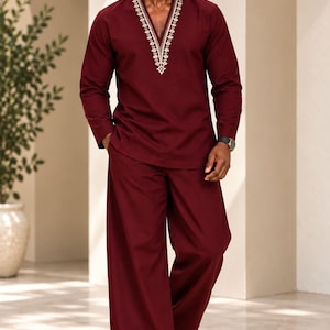 May include: A deep burgundy long-sleeved tunic featuring a V-neckline with intricate white and gold embroidery, paired with matching wide-leg trousers. The ensemble is worn with brown leather sandals and dark-tinted sunglasses.