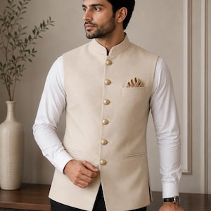 May include: A beige Nehru vest featuring a mandarin collar, six gold-toned metallic buttons down the front, and a matching pocket square in the chest pocket. The vest is worn over a crisp white long-sleeved button-down shirt and black trousers.