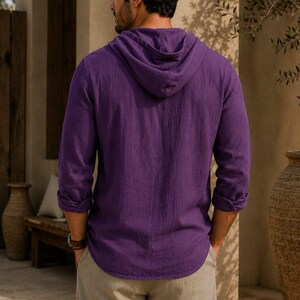 May include: A long-sleeved purple hooded shirt with a textured fabric finish, featuring a hood, cuffed sleeves, and a curved hemline, paired with light beige trousers. The shirt is shown from the back, displaying the hood and sleeve details.