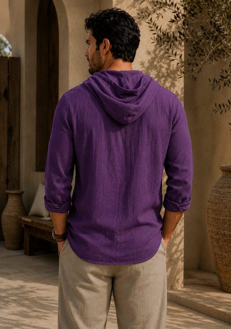 May include: A long-sleeved purple hooded shirt with a textured fabric finish, featuring a hood, cuffed sleeves, and a curved hemline, paired with light beige trousers. The shirt is shown from the back, displaying the hood and sleeve details.