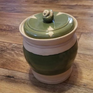 Puede incluir: Un tarro de cerámica verde y blanco con tapa. La tapa tiene un remolino decorativo en la parte superior.
