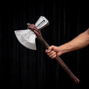 May include: A silver-colored battle axe with a wide, metallic blade featuring intricate geometric engravings. The handle is wrapped in a dark brown, textured material that resembles twisted leather, providing a grip along the shaft.