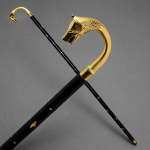 May include: A black walking stick featuring a polished gold-coloured handle shaped like a wolf head. The shaft is decorated with small gold-coloured accents along its length. A second, thinner black walking stick with a smaller wolf head handle is visible.