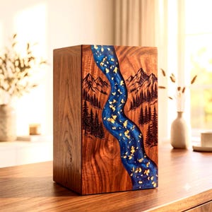 May include: A rectangular wooden urn featuring a central vertical river design made of blue resin with embedded gold leaf flakes. The wood grain is visible on the sides, and the front panel displays a laser-engraved mountain and forest landscape.
