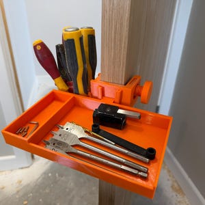 May include: A bright orange 3D-printed clamp-on tool tray attached to a wooden post. The tray features a vertical holder for screwdrivers and a flat section containing three spade drill bits, a small black metal block, and a few small screws.