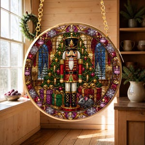 May include: A circular hanging ornament featuring a stained glass style illustration of a nutcracker standing before a decorated Christmas tree with wrapped gifts and a small mouse, framed by a gold border and suspended by a gold chain.