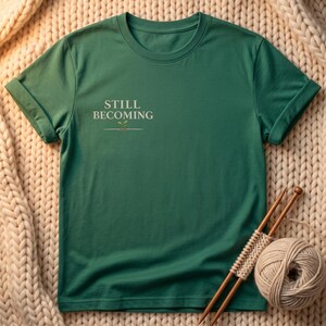 May include: A forest green short-sleeved t-shirt featuring the text "STILL BECOMING" printed in a light cream color on the upper left chest, accompanied by a small graphic of a tiny green sprout growing from a thin horizontal line.