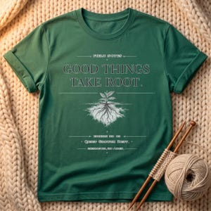 May include: A forest green short-sleeved t-shirt featuring white text that reads FIELD NOTES, GOOD THINGS TAKE ROOT, EDITION NO 05, QUIET GROWTH DEPT, and CATALOG NO, 134 / ARCH, centered above a detailed white graphic of a plant with roots.
