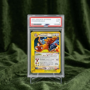May include: A graded 2023 Pokemon Skyridge Charizard-Holo trading card encased in a clear plastic slab. The card features a yellow border, an illustration of the orange Charizard creature, and text including 110 HP, Fireblast, and Dragon Tail attacks.