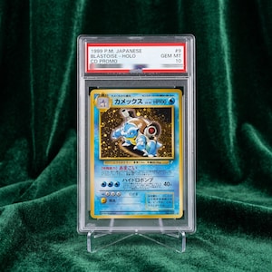 May include: A 1999 Japanese Blastoise holographic trading card encased in a clear protective plastic slab. The label at the top reads 1999 P.M. JAPANESE BLASTOISE - HOLO CD PROMO #9 GEM MT 10. The card features a blue character with cannons.
