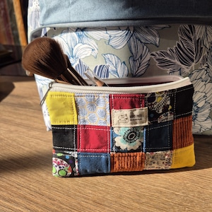 Peut inclure: Une pochette rectangulaire à fermeture éclair composée de carrés de tissu en patchwork de différentes couleurs et motifs, dont jaune, rouge, bleu et noir, avec une étiquette "HAND MADE Natural Style" et un pinceau de maquillage dépassant.