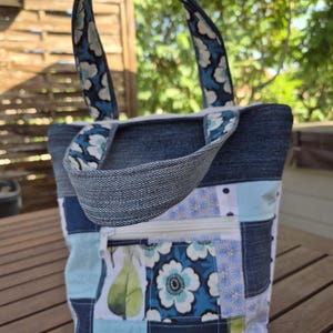 Peut inclure: Un sac fourre-tout en patchwork composé de denim bleu et de divers panneaux de tissu à motifs floraux dans des tons de bleu, blanc et vert. Le sac comprend une poche frontale zippée et deux anses en denim assorties avec une doublure florale.