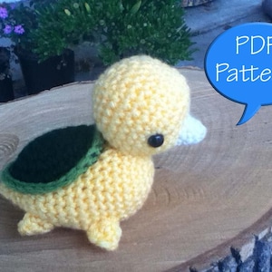Peut inclure: Un canard jaune en crochet avec une carapace verte sur le dos. Le canard a des yeux noirs et un bec blanc. Le canard est assis sur une surface en bois. Le texte "PDF Pattern!" est dans une bulle de dialogue bleue.